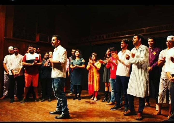 A theatre ensemble gathered on a warmly lit stage, with a man speaking into a microphone and performers applauding